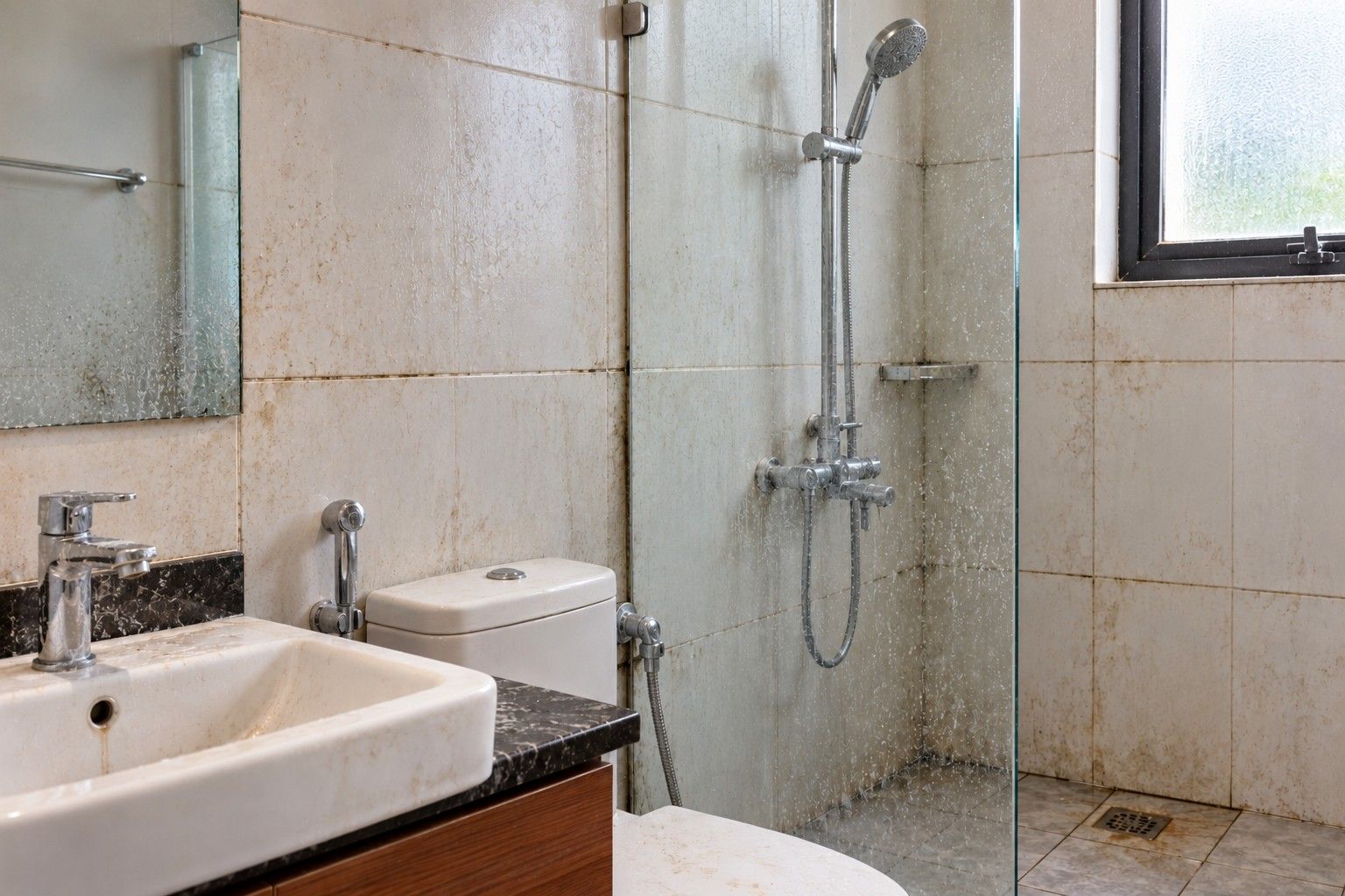 A bathroom wall and fittings with limescale, soap scum and dark grout before deep cleaning